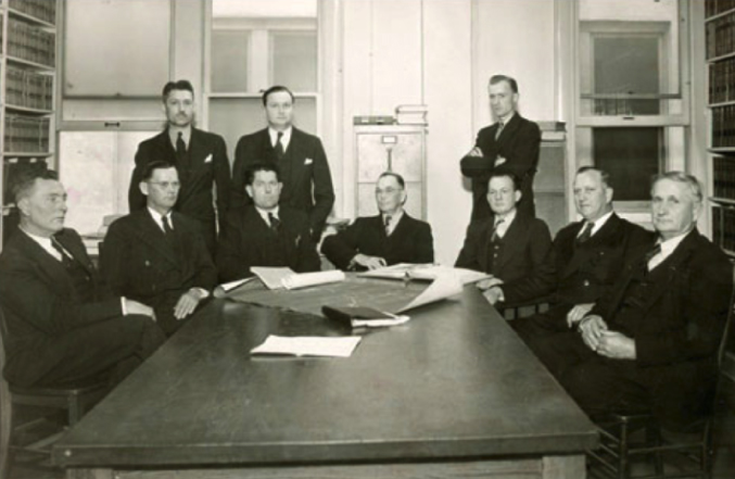 A black and white historical photo of board members from Satilla EMC seated at a table. Seven men are seated and three are standing behind the group. All men are wearing suits and ties.