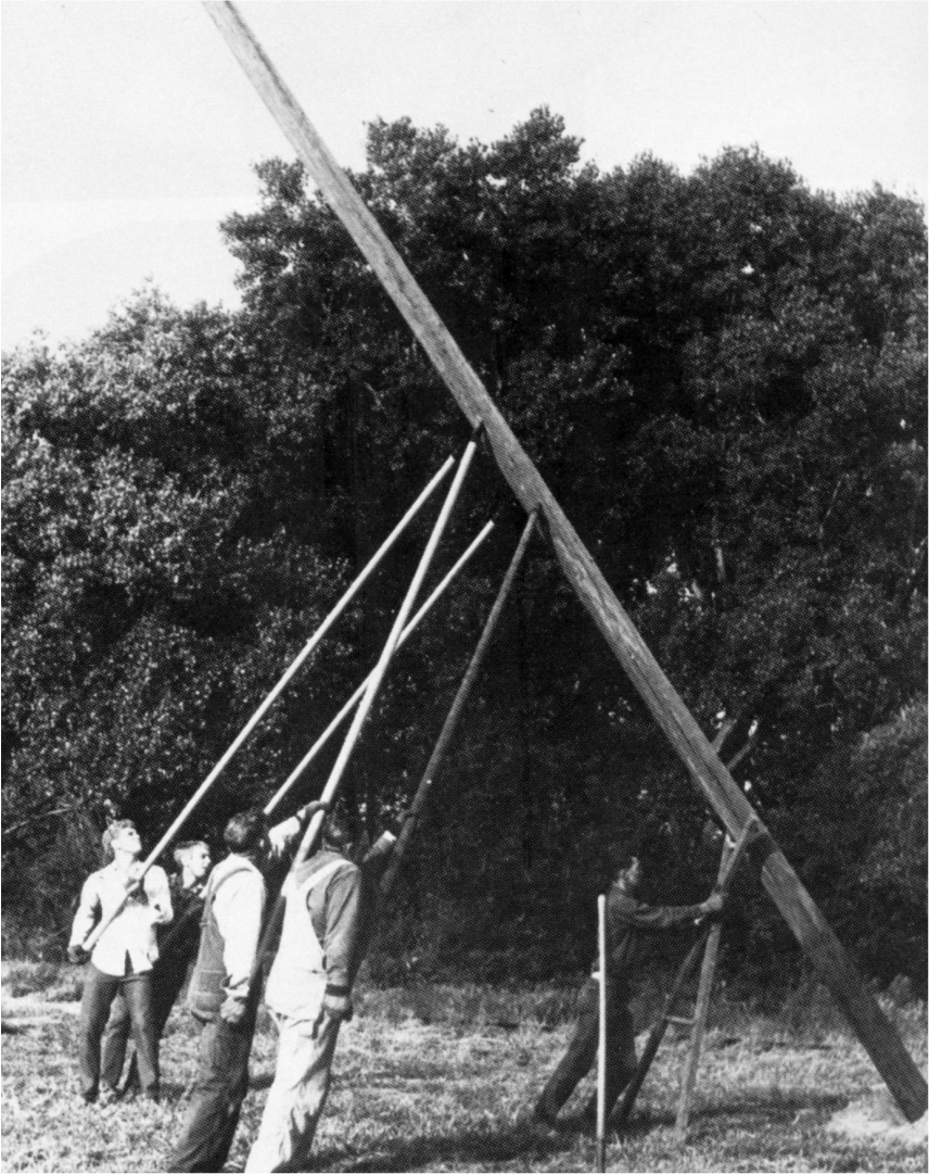 A black and white photo of men raising an electric pole by hand with the use of longer poles. The electric pole leans at an angle as they raise it.